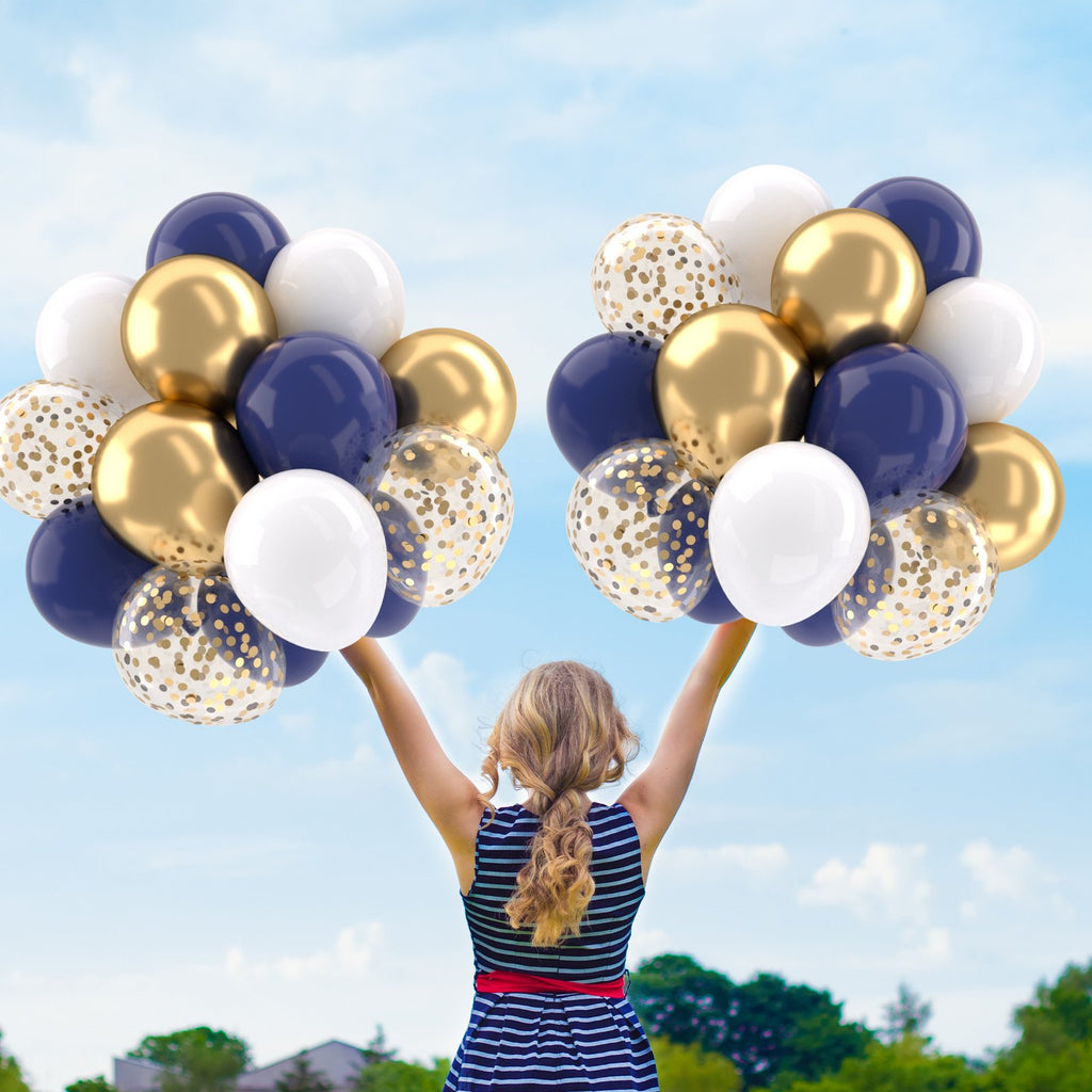 Woman holding navy blue, gold, white, and confetti balloons against a blue sky