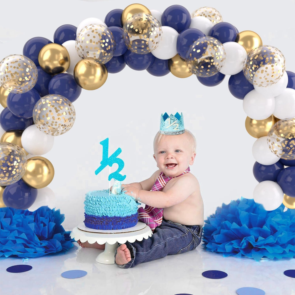 Baby wearing a crown and tie sits with a cake, surrounded by a balloon arch and blue tissue paper decorations.