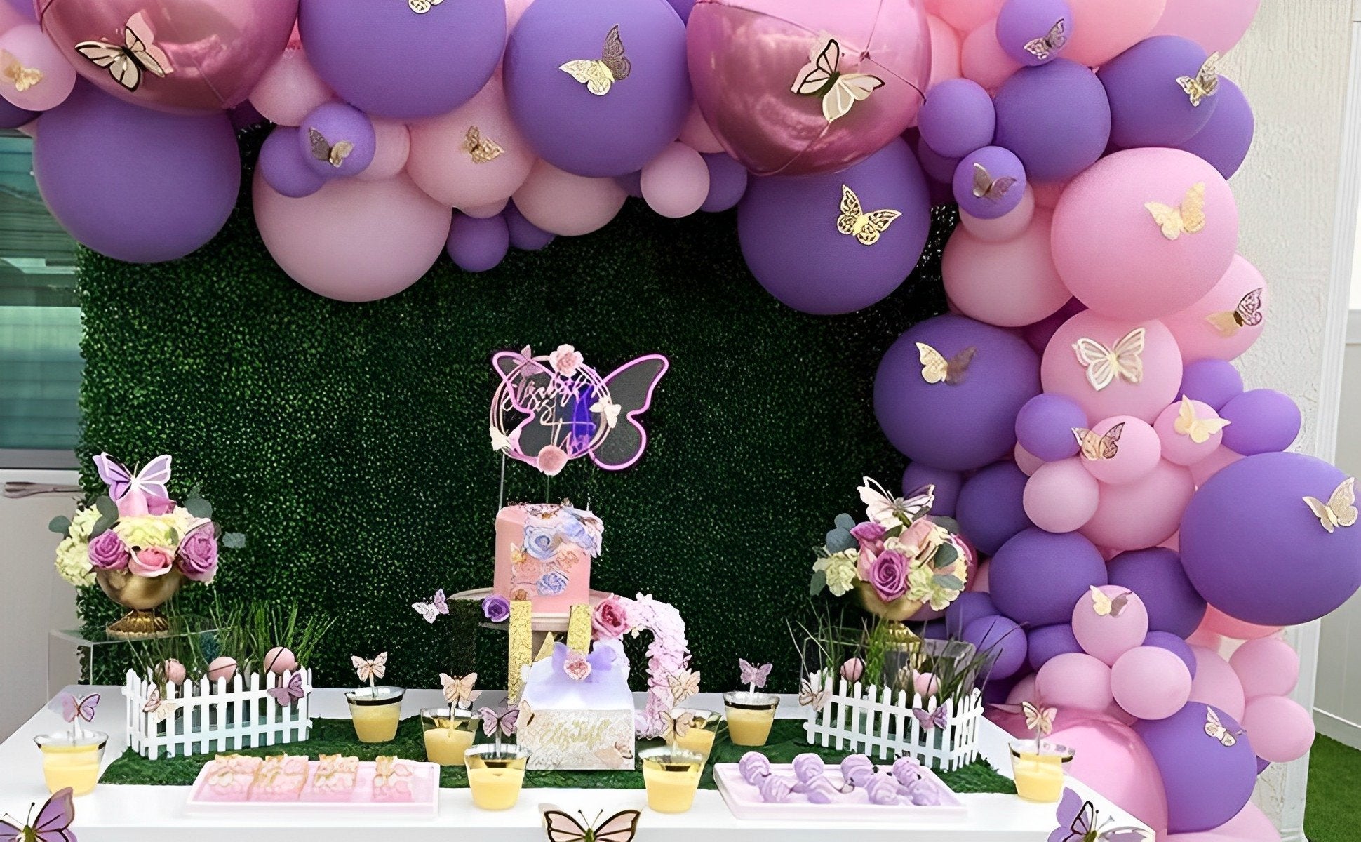 Party table with pink and purple balloons, cake, flowers, and desserts, decorated with butterfly accents.