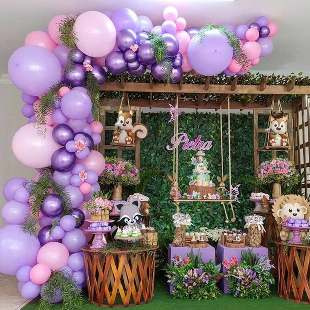 Forest-themed party decor with pink and purple balloon arch, cake on a swing, and woodland creature decorations.