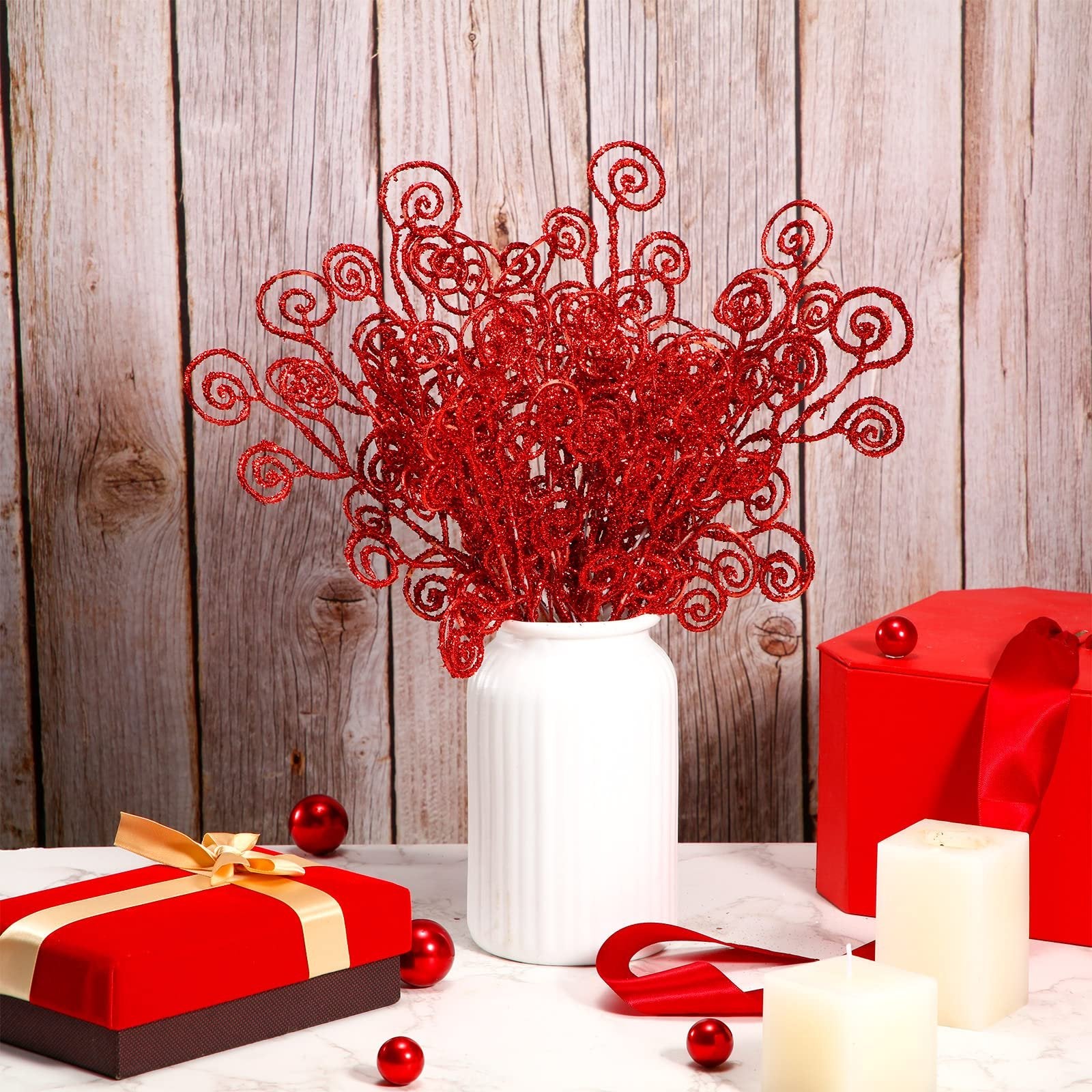 Red glitter curly twig picks in a white vase with Christmas decor, including gift boxes and candles.
