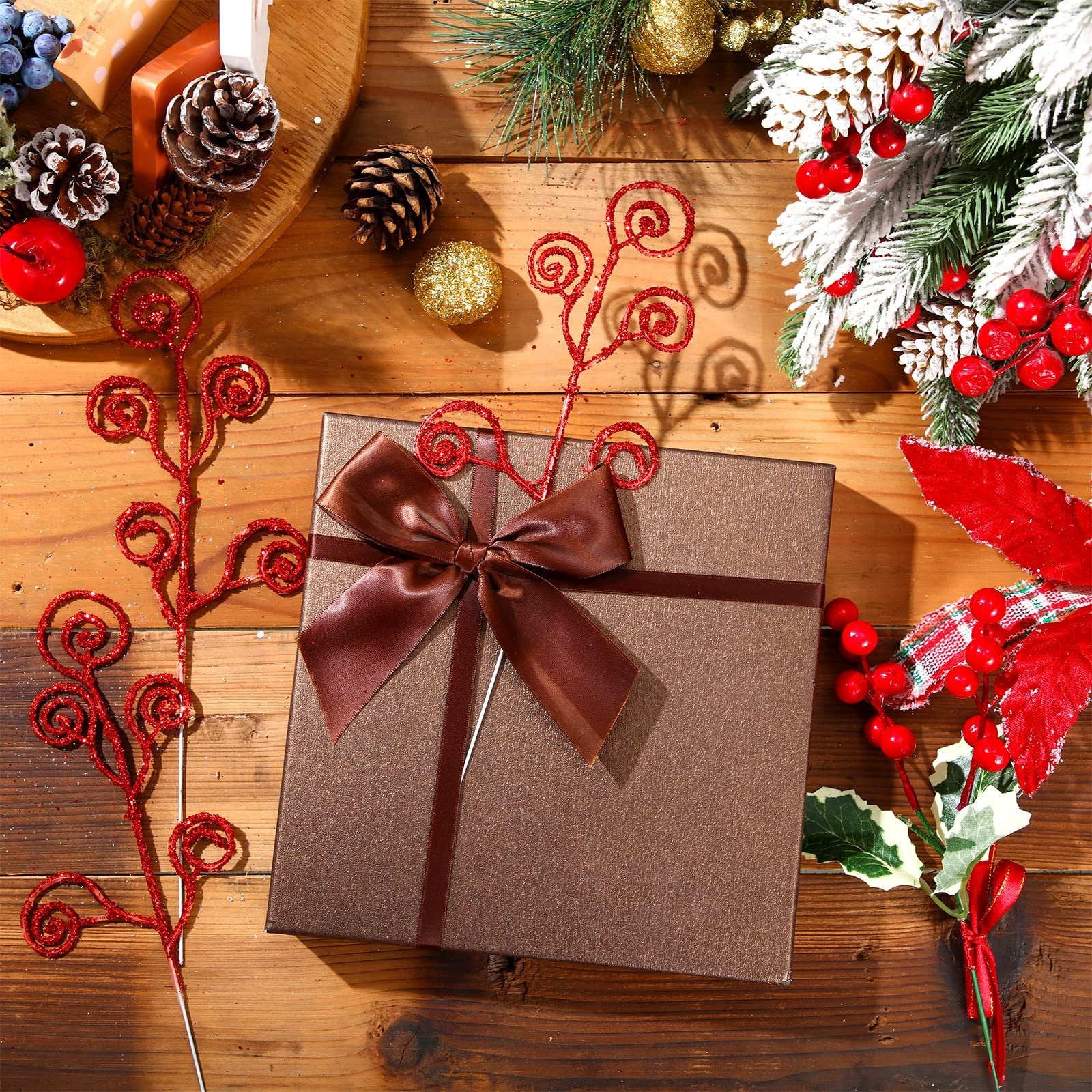 Christmas gift box with a brown ribbon bow surrounded by festive decorations including glittery red branches, pinecones, and frosted greenery.