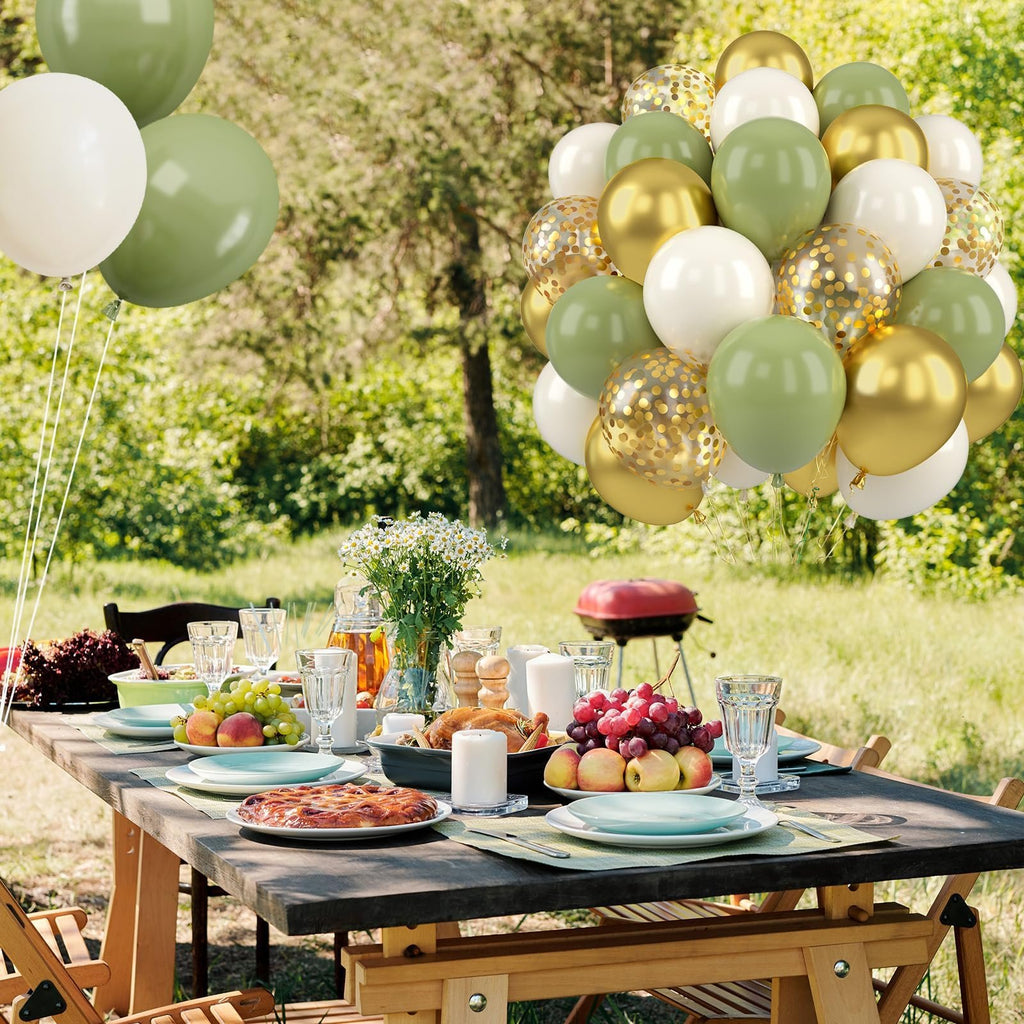 A cluster of sage green, gold, and white balloons with confetti floats above a picnic table set for a meal outdoors.