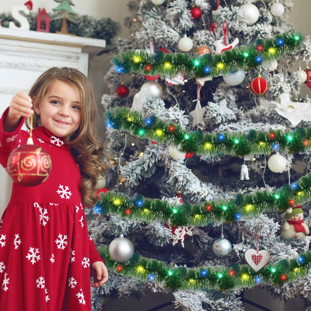 Young girl in a red snowflake dress holding a Christmas ornament in front of a decorated tree with tinsel garland and lights