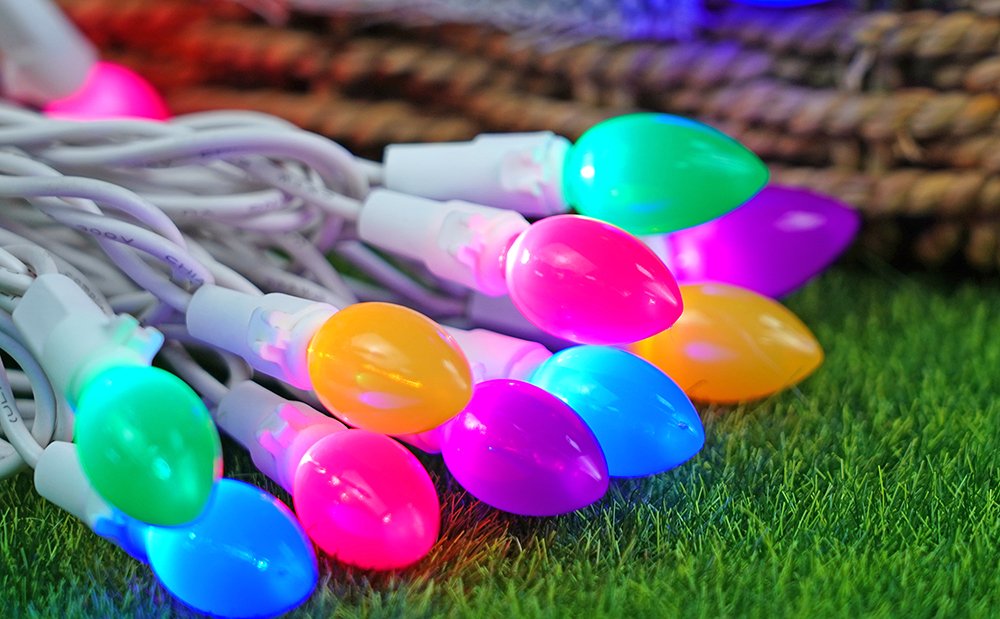 Close-up of multicolor LED string lights with white wires on green grass and a rope background.