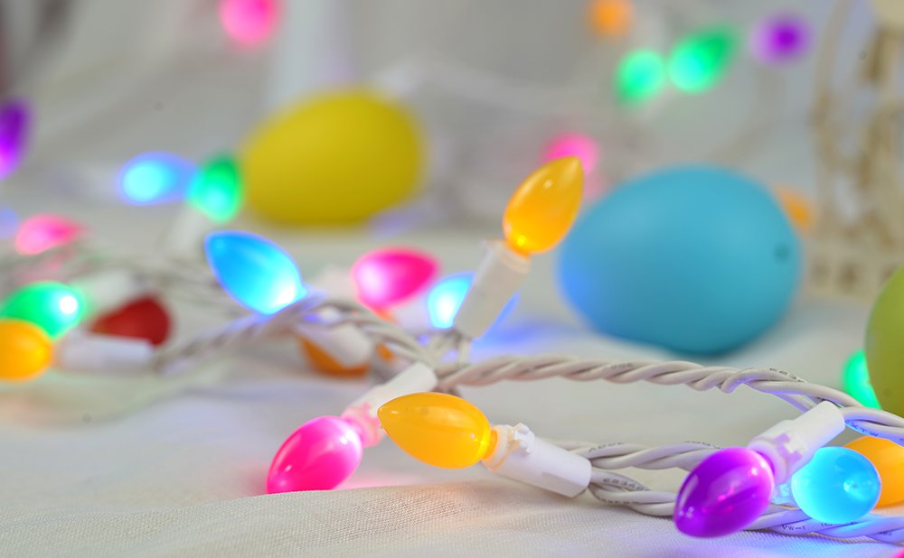 Multicolor Easter egg string lights with white wire on a textured white surface, with blurred Easter eggs in the background.