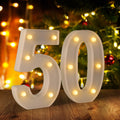 White marquee numbers 5 and 0 with illuminated bulbs, set on a wooden surface with a blurred Christmas tree and bokeh lights in the background.