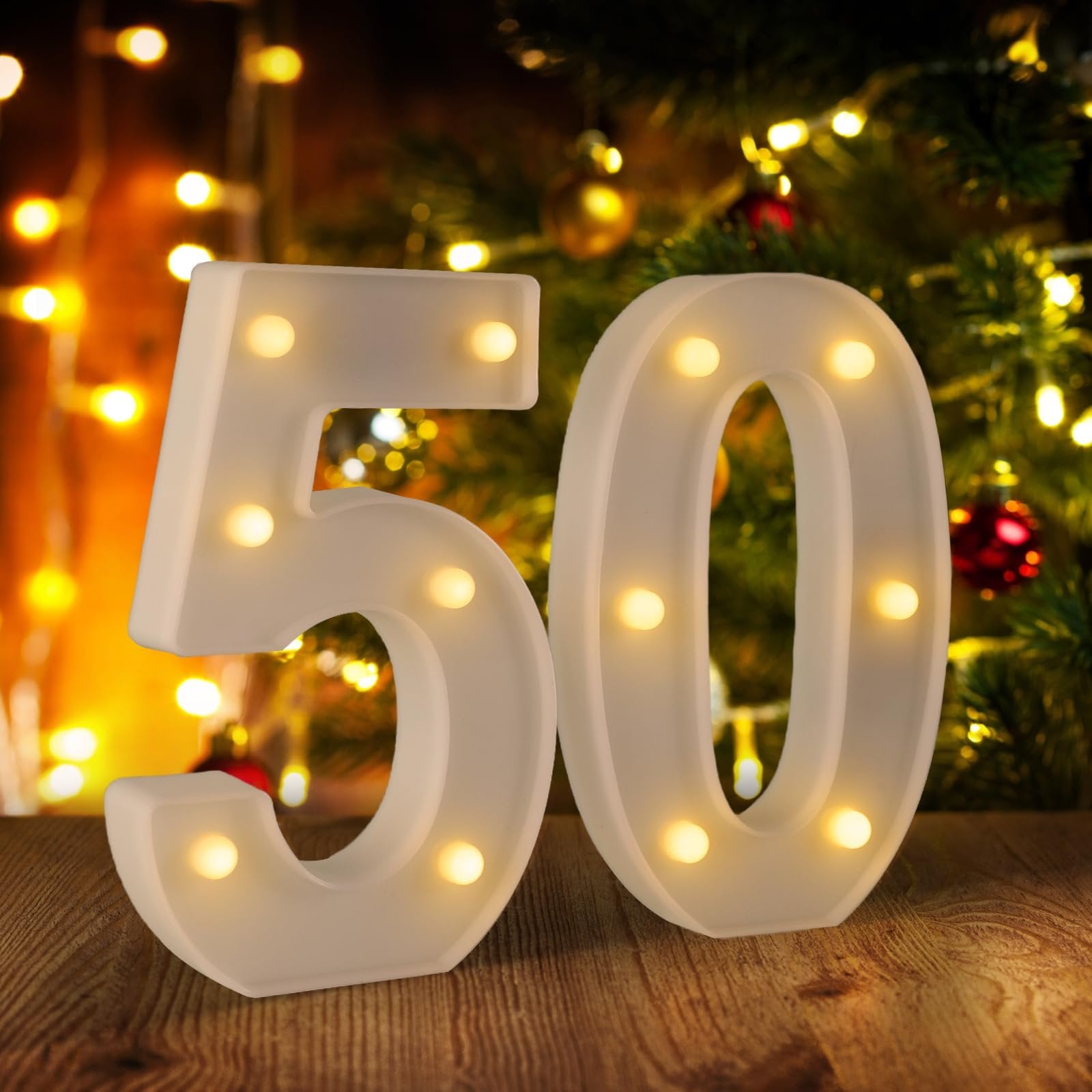 White marquee numbers 5 and 0 with illuminated bulbs, set on a wooden surface with a blurred Christmas tree and bokeh lights in the background.