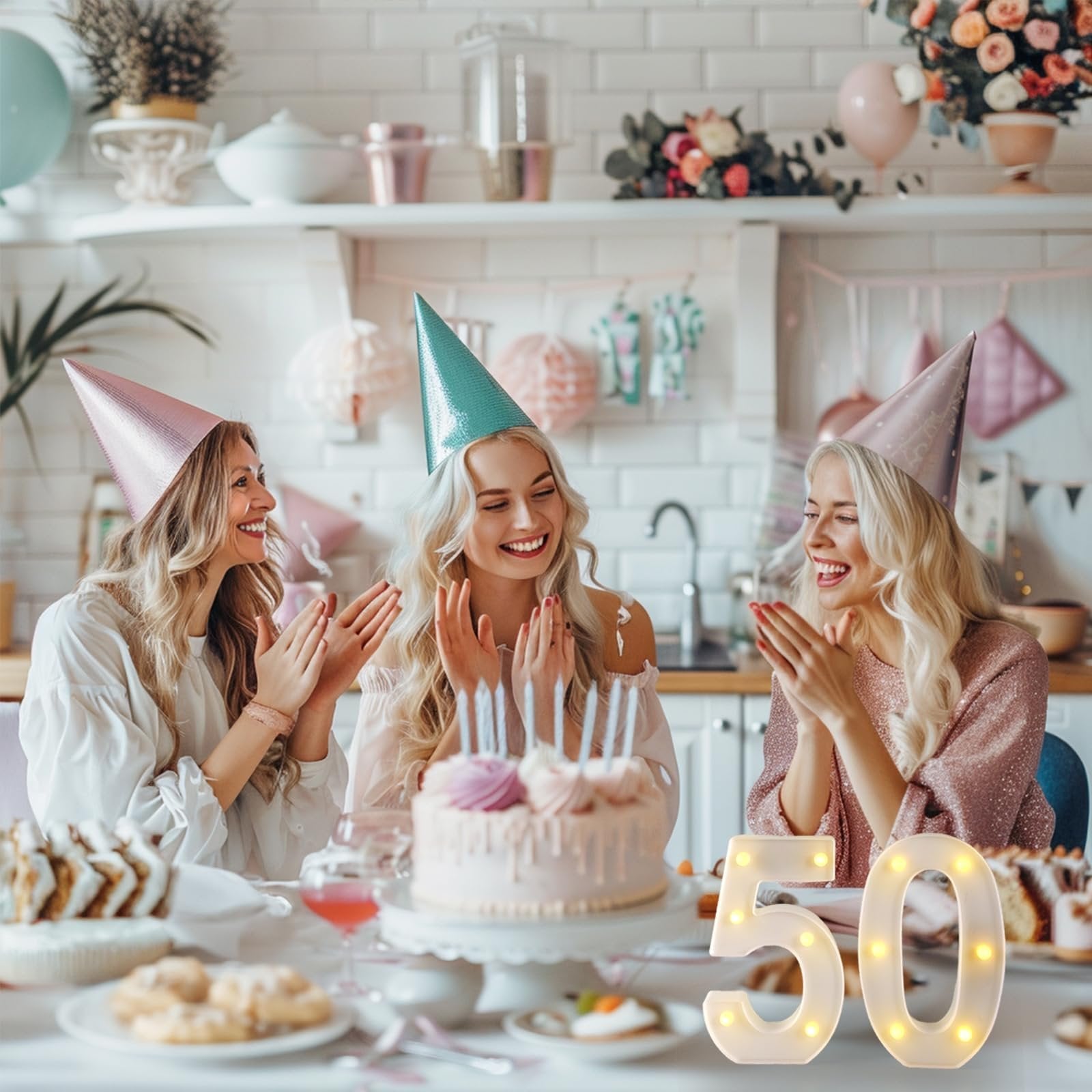 Three women in party hats celebrating a birthday with a cake and marquee numbers '50'.