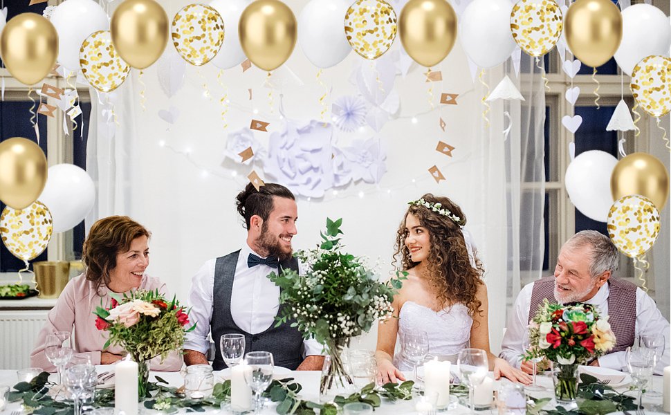 Wedding party seated at a decorated table with gold, white, and confetti balloons overhead.