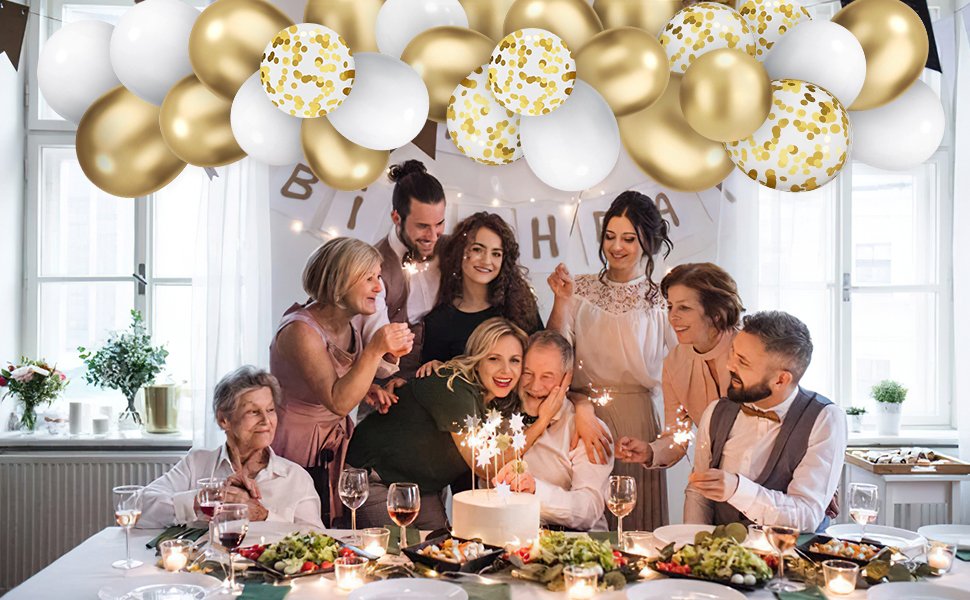 Family celebrating around a table with a cake and sparklers, white and gold balloons overhead