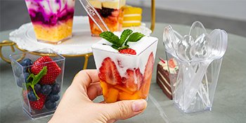 Hand holding a clear plastic dessert cup filled with strawberries and mango, topped with mint. Other cups with fruit and desserts are visible.
