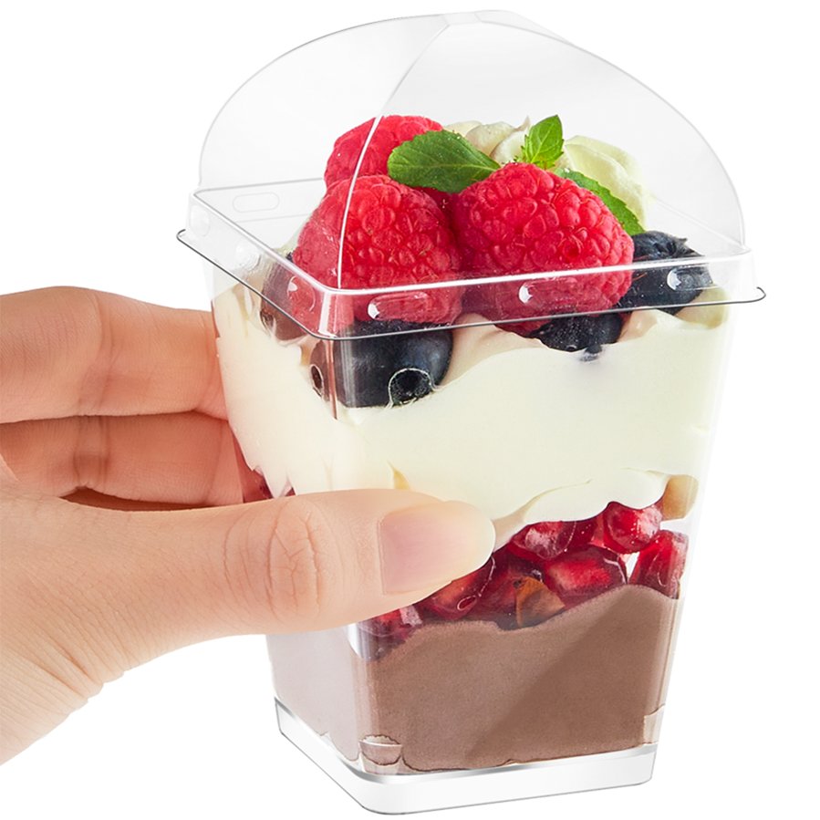 Hand holding a clear plastic dessert cup filled with layered chocolate mousse, cream, raspberries, blueberries, pomegranate seeds, and mint.