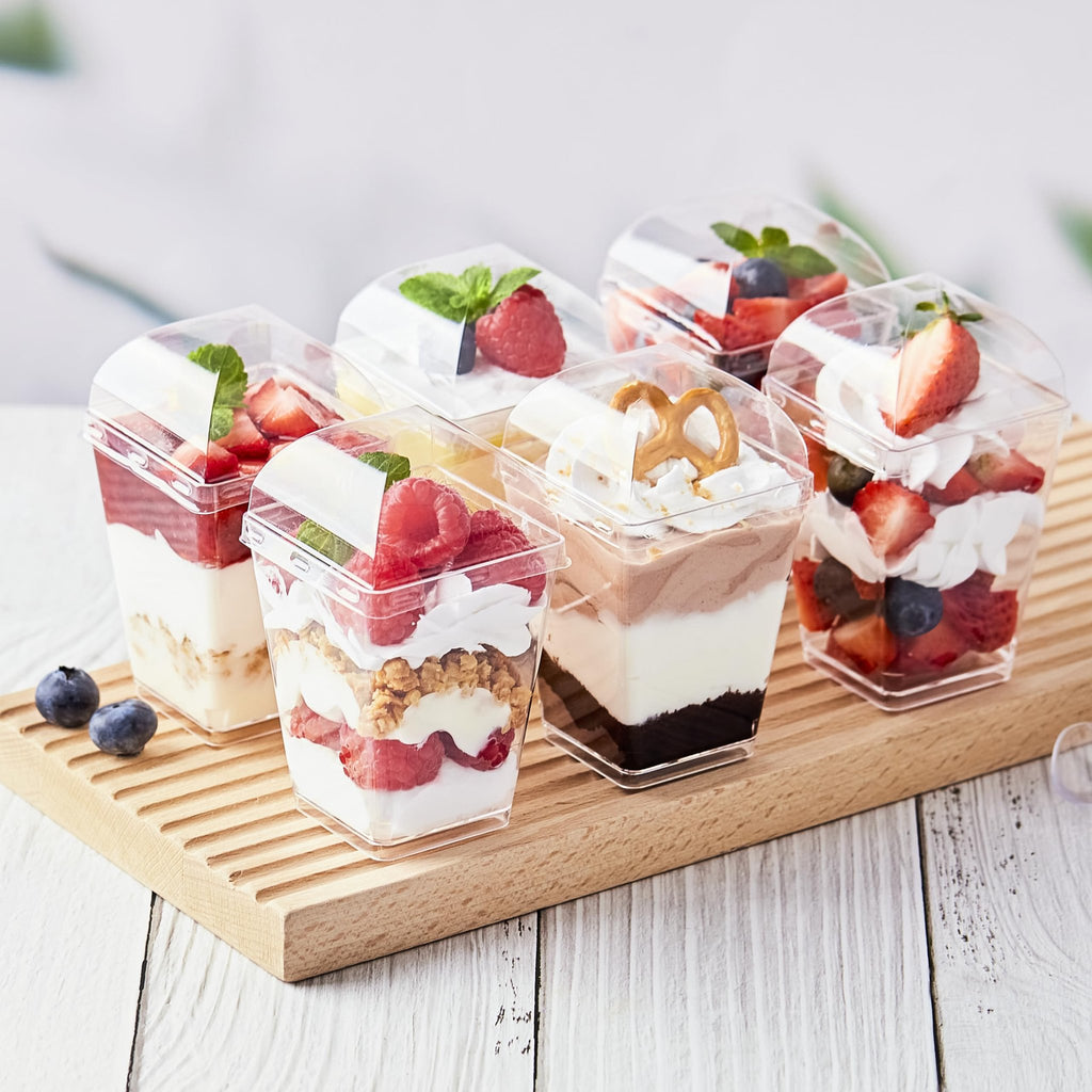 Six clear plastic dessert cups filled with layered desserts and topped with fruit and mint, arranged on a wooden board.