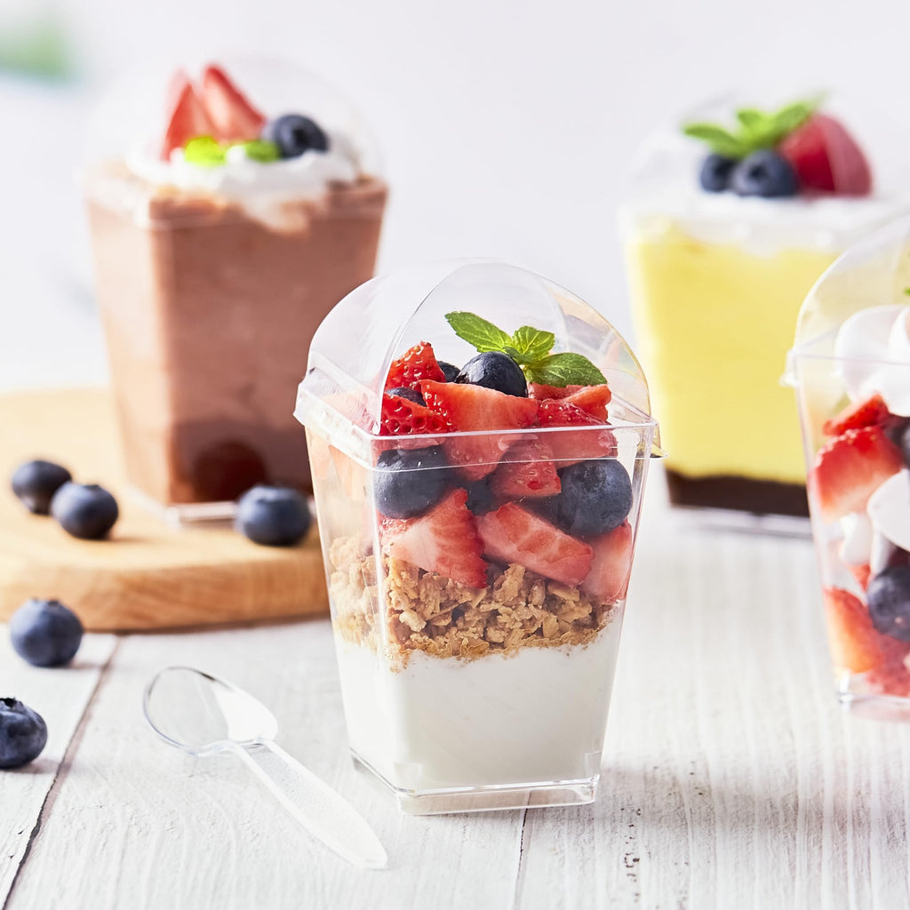 Clear plastic dessert cups filled with yogurt, granola, strawberries, and blueberries, with lids and spoons nearby.