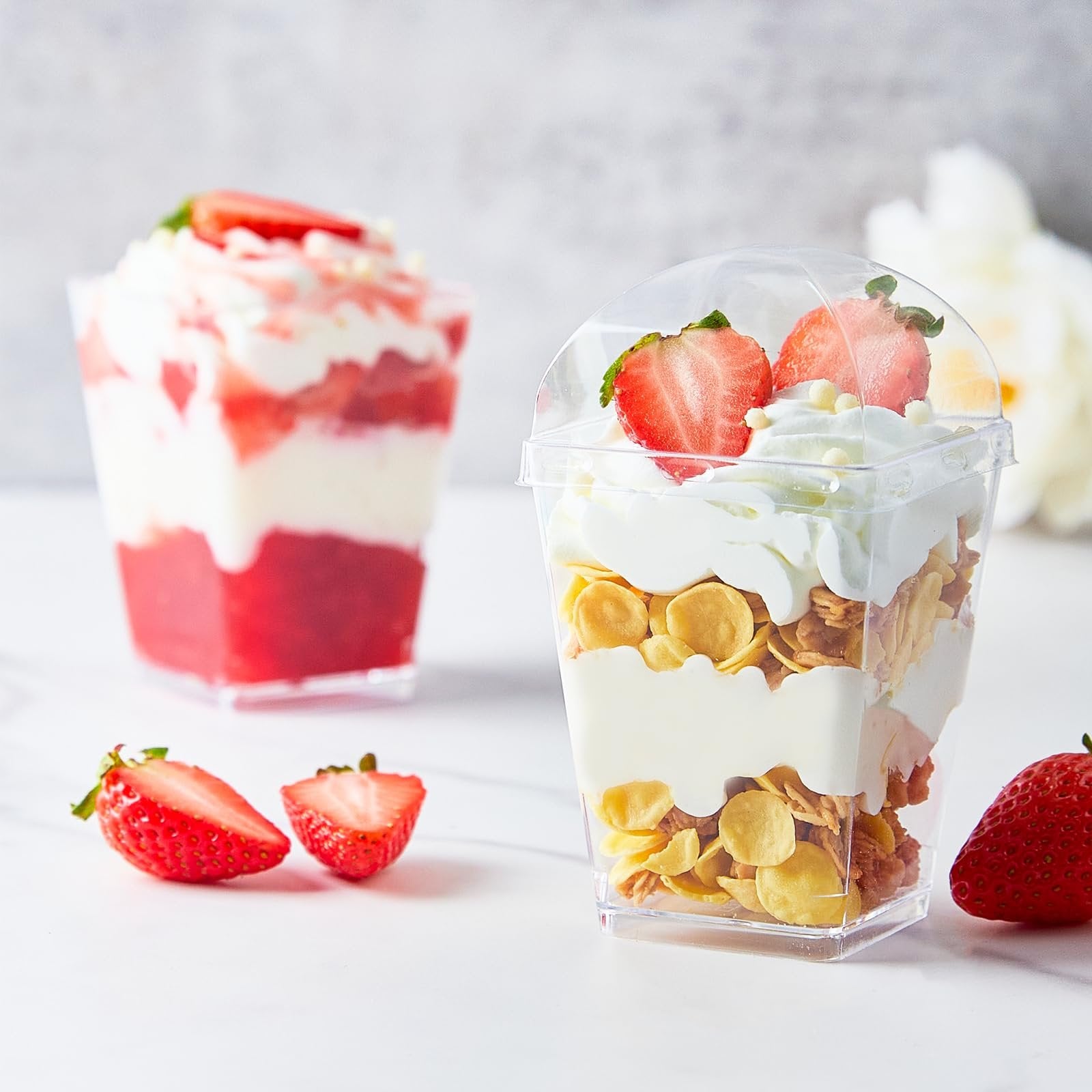 Two clear plastic dessert cups filled with layered yogurt, granola, and strawberries, with fresh strawberries on the side.
