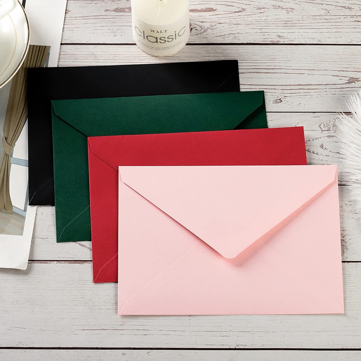 Four colorful envelopes (pink, red, dark green, black) fanned out on a wooden surface with a candle and decorative feather.