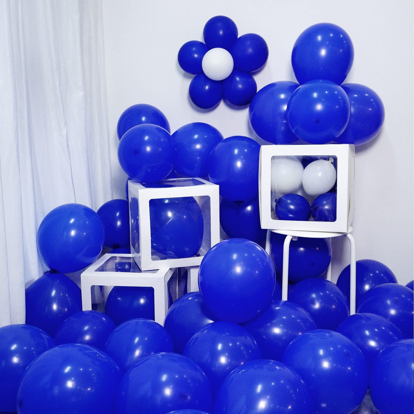 Pile of royal blue latex balloons with clear display boxes and a white balloon flower decoration on a white wall.