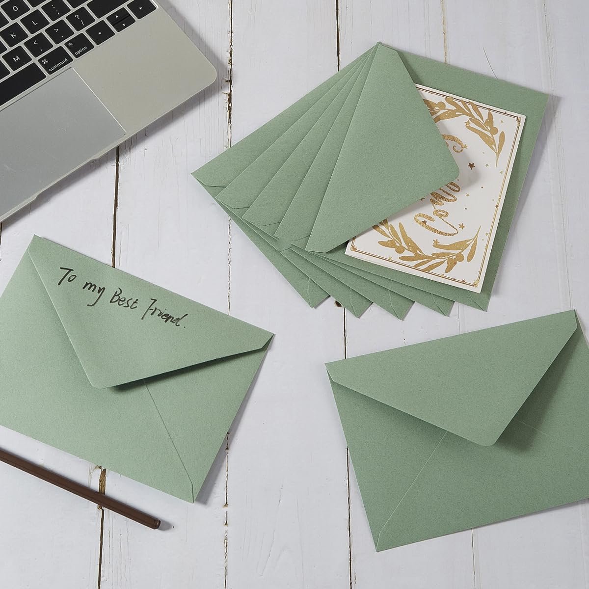 Sage-green A7 envelopes and a card with gold floral design, arranged on a white wooden surface with a laptop keyboard and a pencil.
