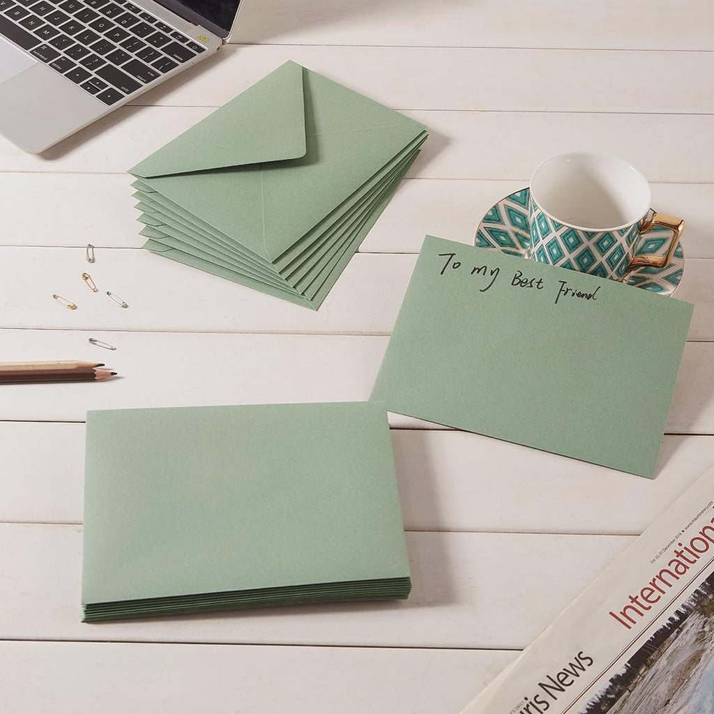 Sage-green A7 envelopes stacked on a white wooden desk with a laptop, cup, and newspaper.