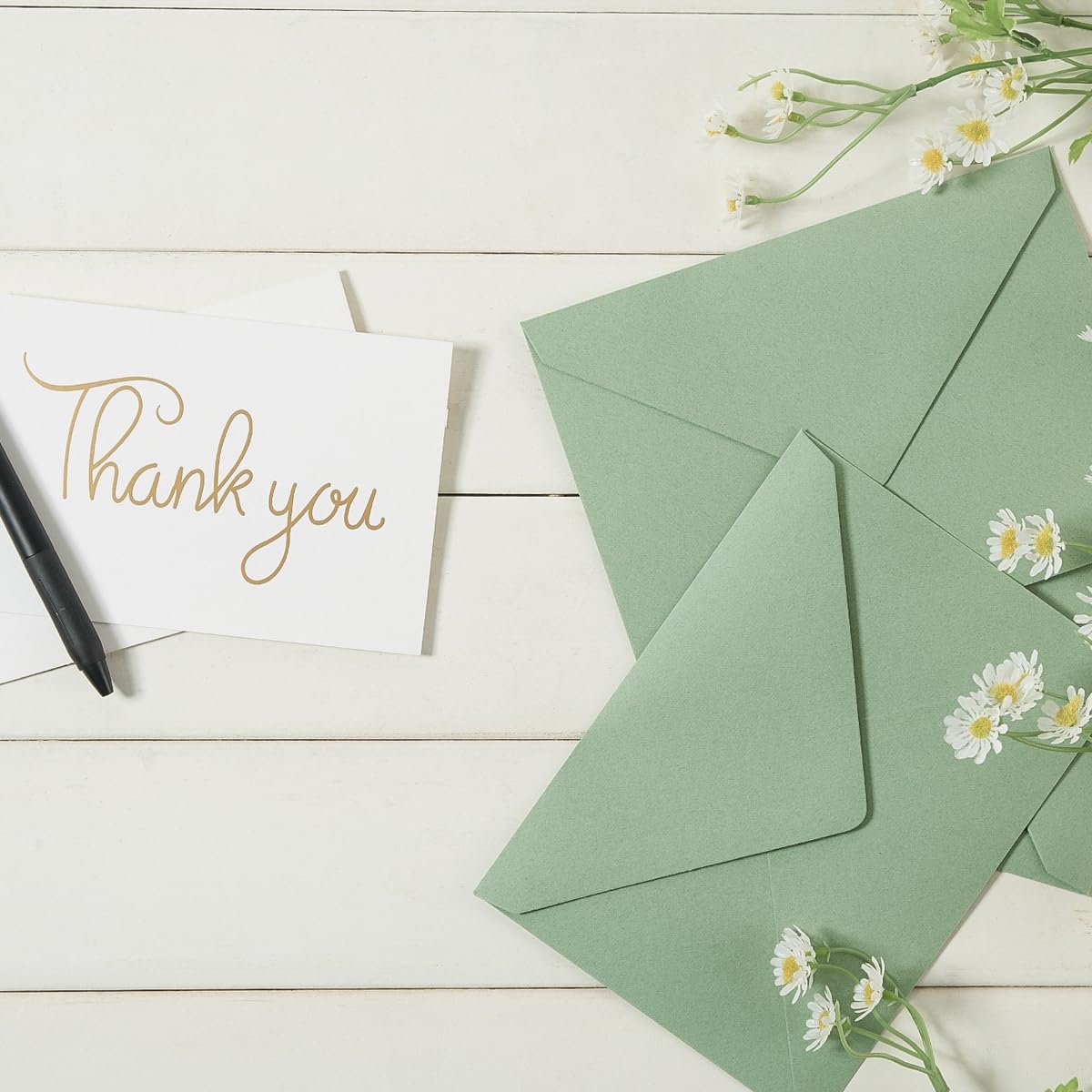 Sage green A7 envelopes and a 'Thank You' card with a pen on a white wooden surface, decorated with small white flowers.
