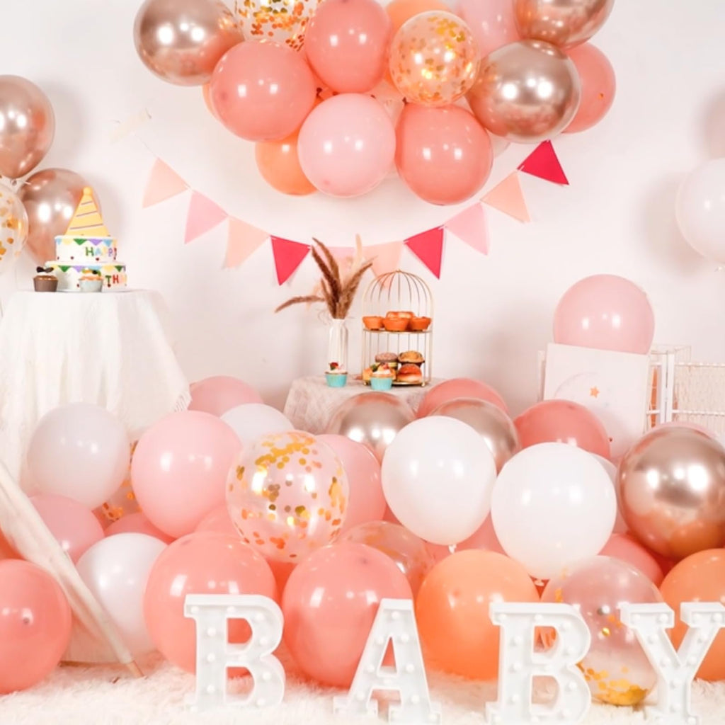A festive arrangement of pink, gold, and white balloons with confetti, a cake, and decorative letters spelling 'BABY'.