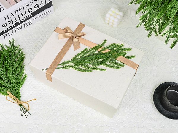 White gift box with gold ribbon and faux pine sprigs, next to a book and decorative candle.