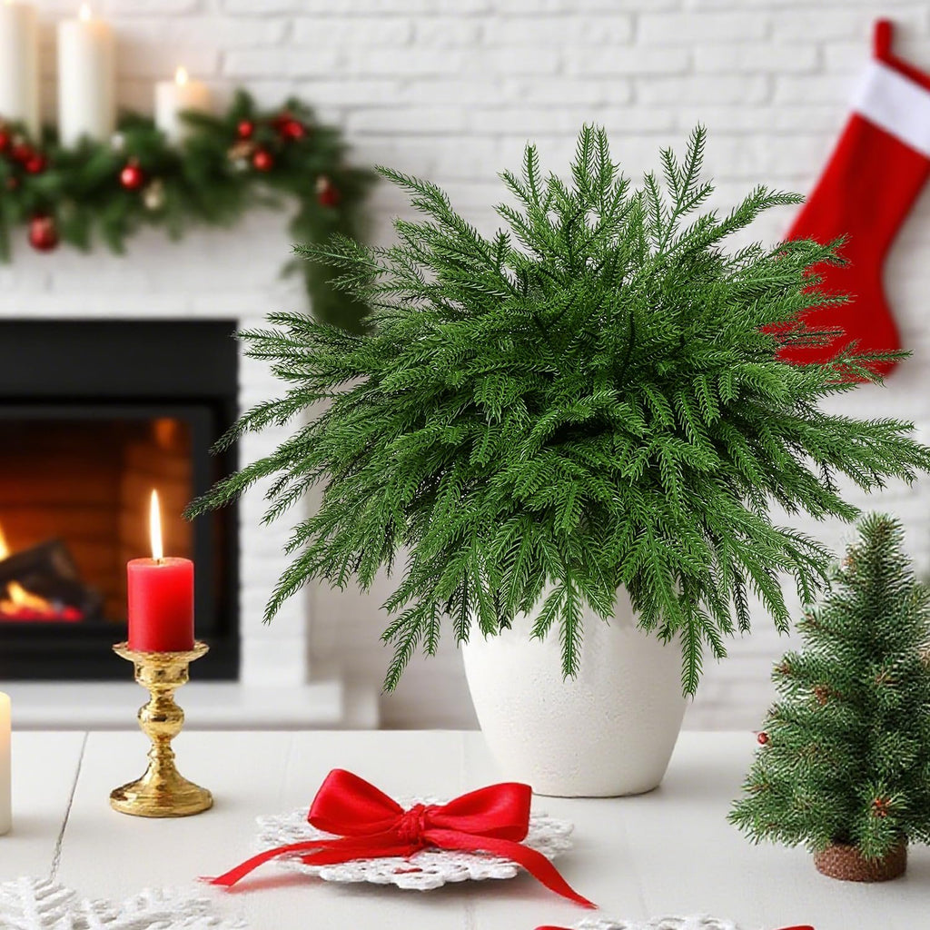 Artificial Norfolk pine stems in a white pot on a table with Christmas decorations, including a fireplace and stocking.