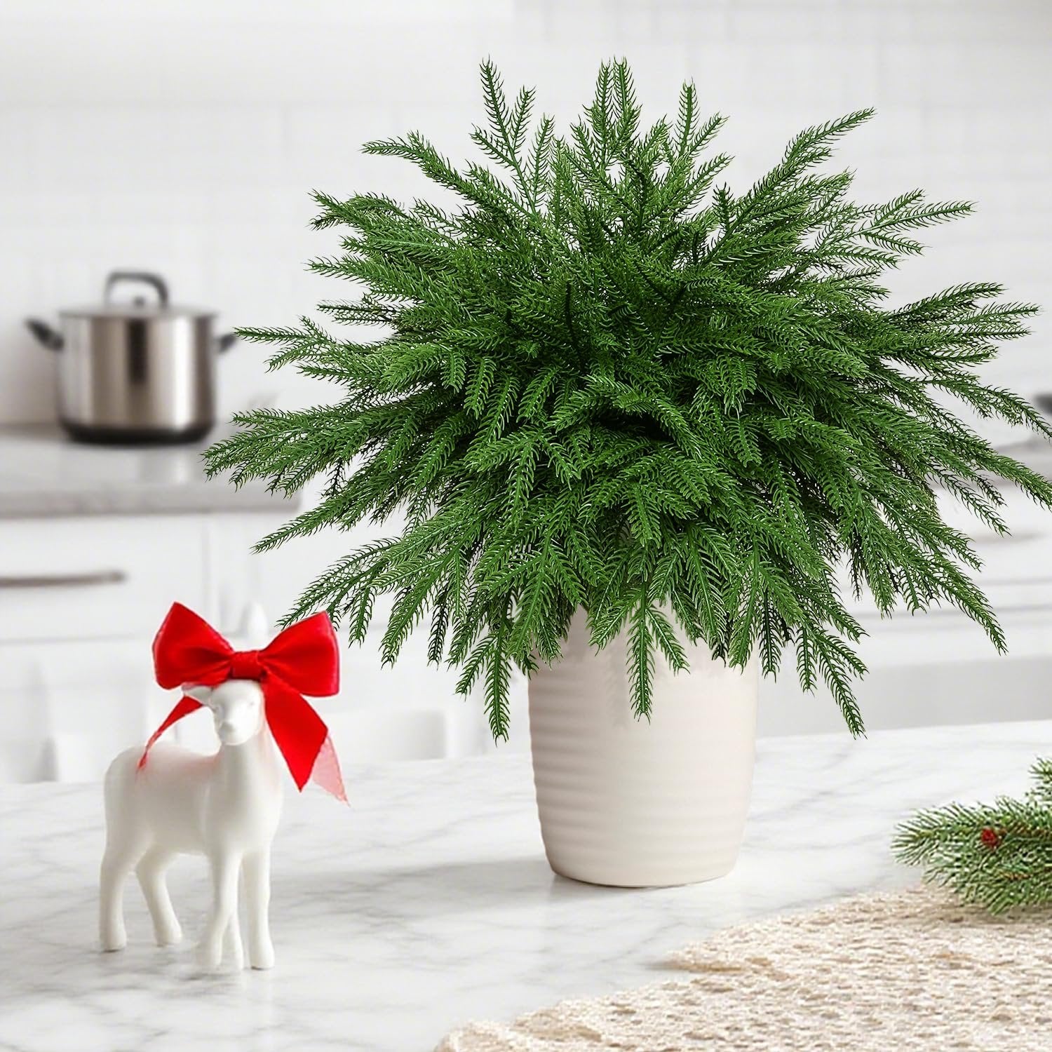 Artificial Norfolk pine stems in a white vase with a decorative reindeer and red bow on a marble surface.
