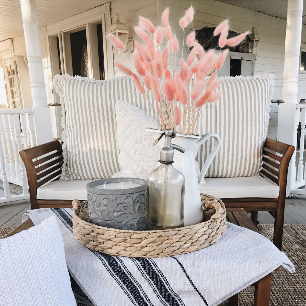 Pink bunny tail grass in a pitcher on a tray with a candle and bottle on a porch bench.