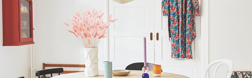 A vase of pink bunny tail grass on a round table with candles and a bowl, next to a red cabinet and floral curtains.
