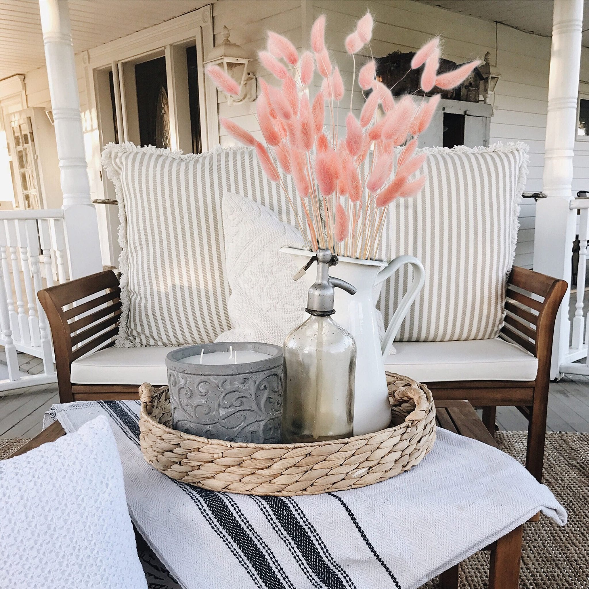 Pink bunny tail grass in a white pitcher on a tray with a candle and vintage bottle on a porch bench