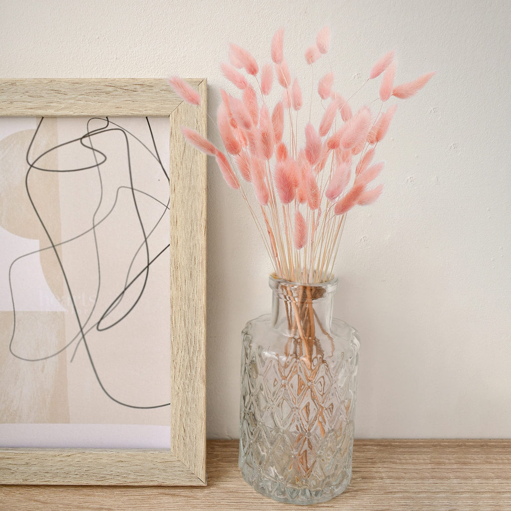 Pink bunny tail grass in a clear glass vase next to a framed abstract art piece on a wooden shelf.