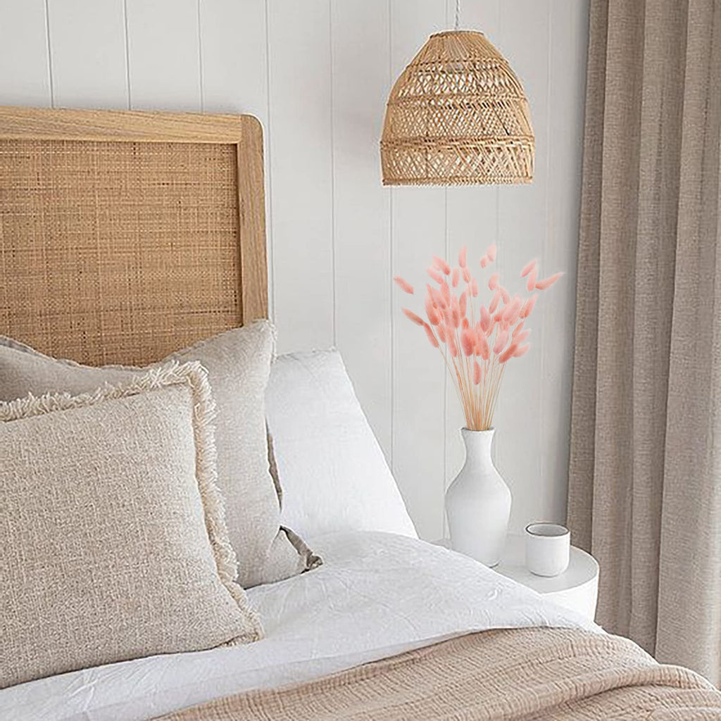 Pink bunny tail grass in a white vase on a nightstand next to a bed with boho-style decor.