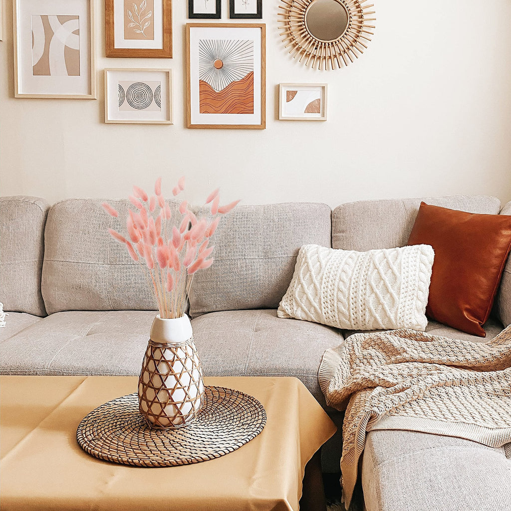 Pink bunny tail grass in a woven vase on a table in front of a sofa with decorative pillows and a blanket.