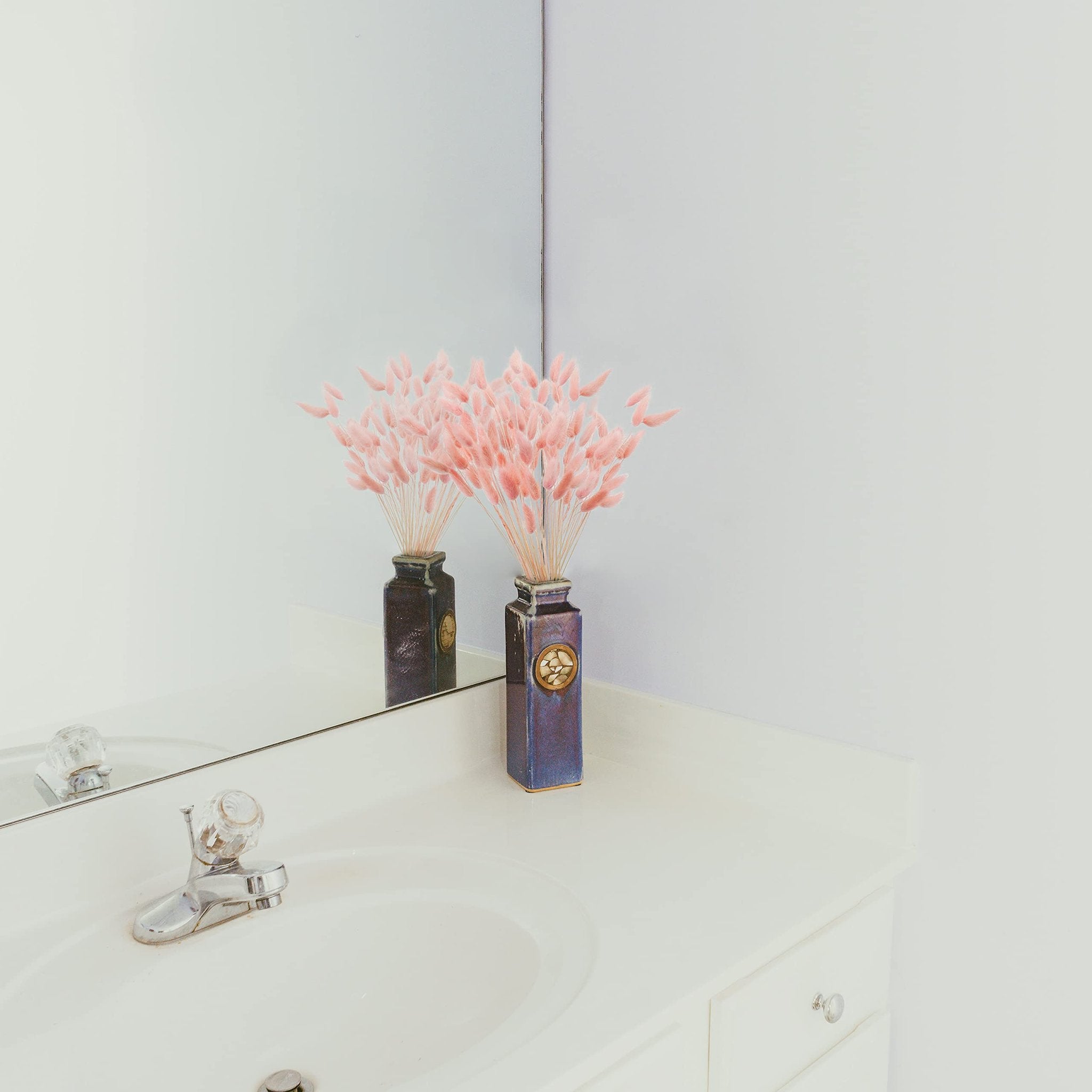 Pink bunny tail grass in a decorative vase on a bathroom counter with a sink and faucet reflected in the mirror.