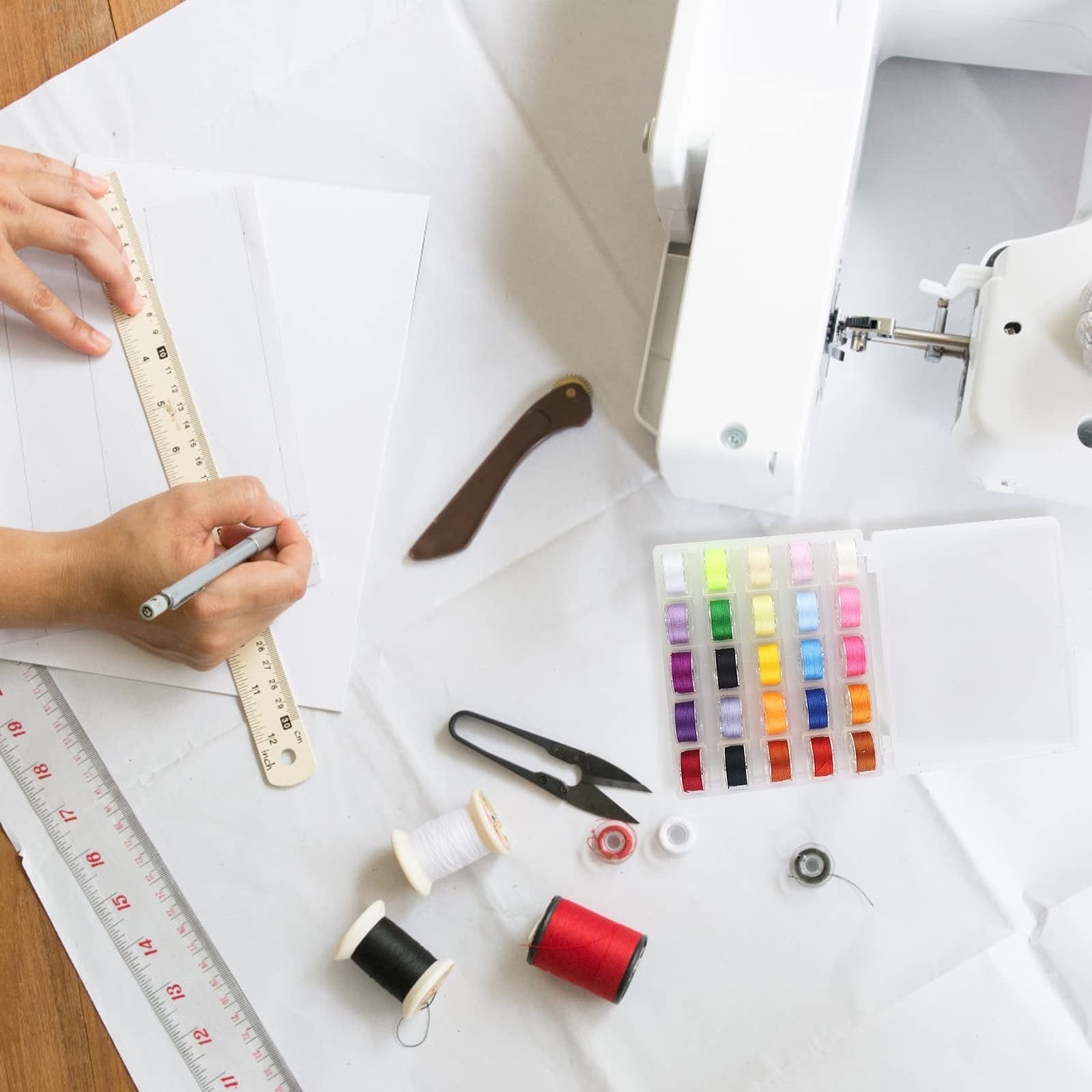 Overhead view of sewing supplies including a sewing machine, colorful bobbins, thread spools, scissors, and rulers on white paper.