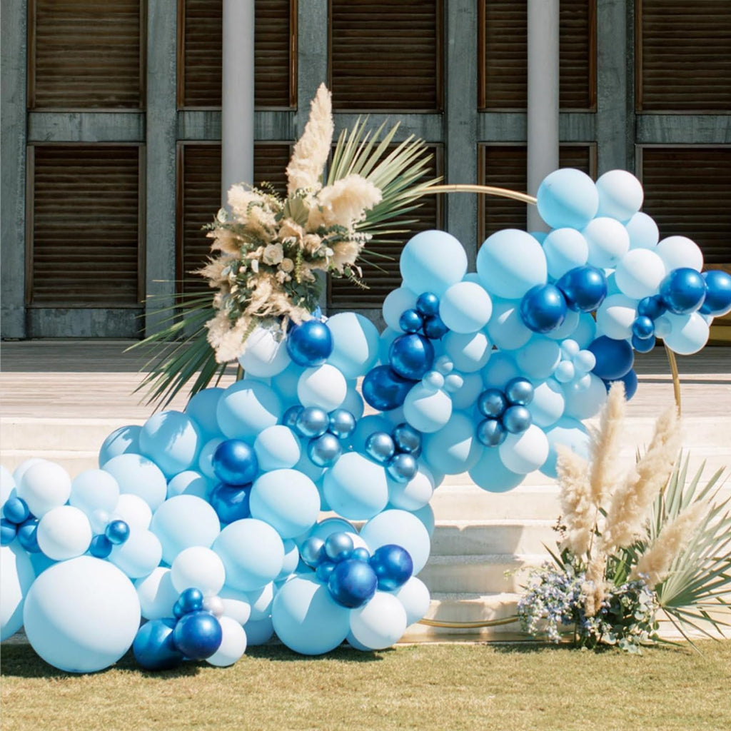 Blue and white balloon arch with metallic accents and dried floral arrangements on grass with stairs and building in background