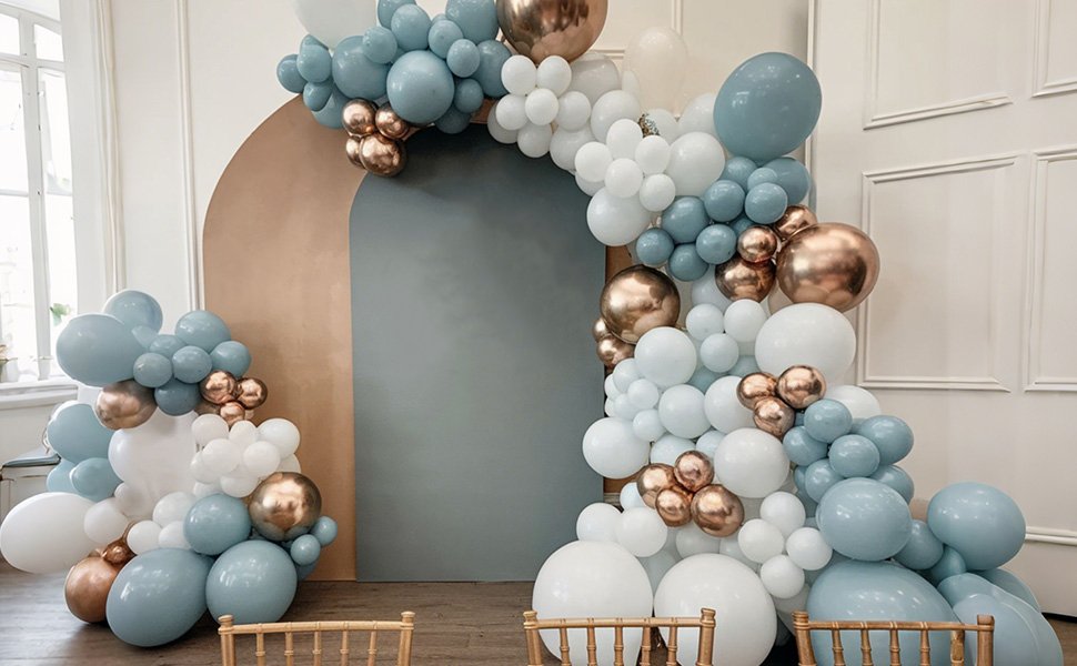 Balloon arch with dusty blue, gold, and white balloons, set against a neutral backdrop with chairs in the foreground.