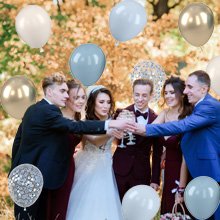 Wedding party toasting with champagne glasses surrounded by dusty blue, gold, and confetti balloons outdoors