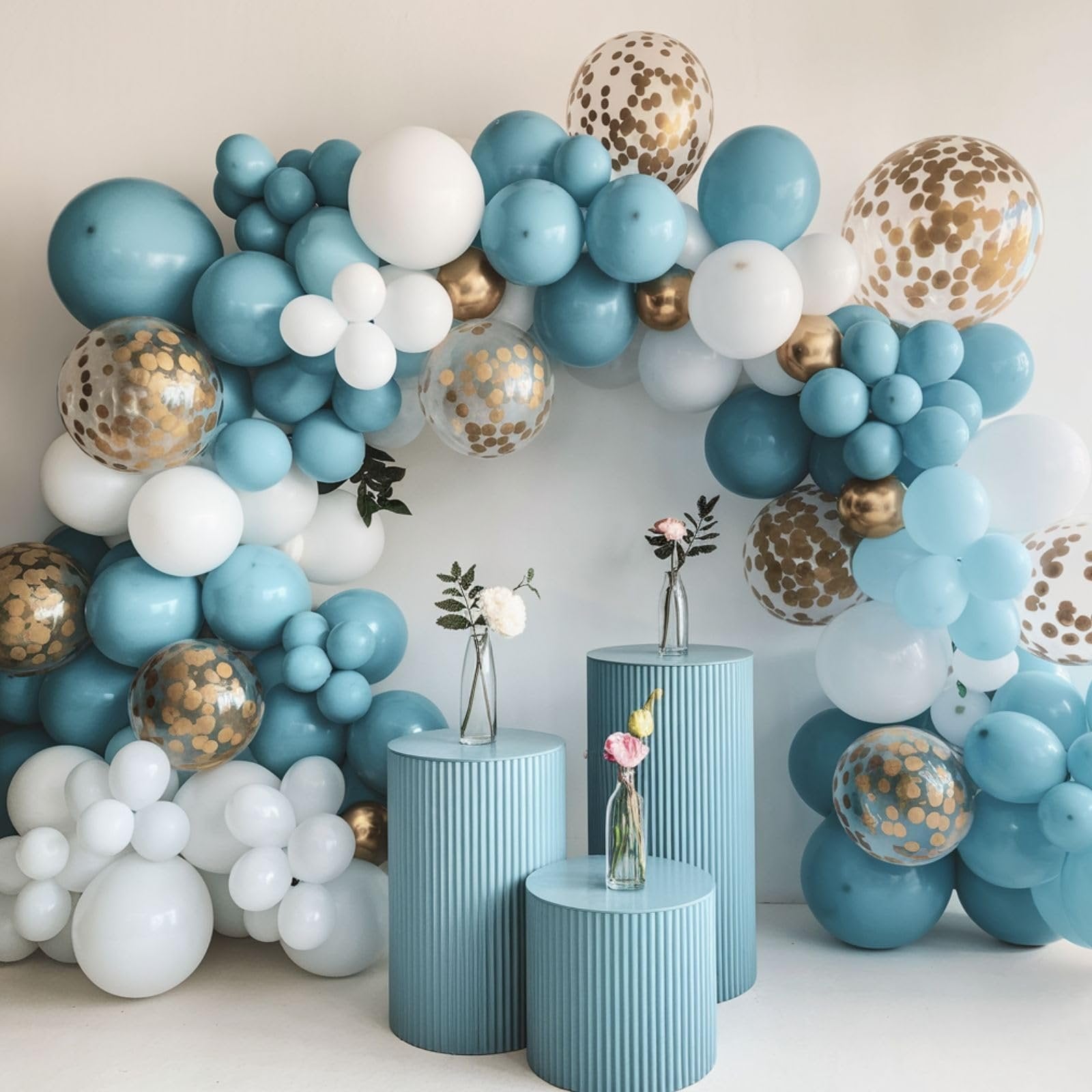 Dusty blue, white, and gold confetti balloons arranged in an arch with decorative pedestals and vases of flowers.