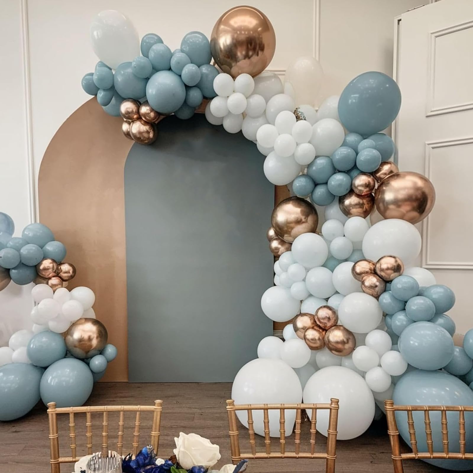 Dusty blue, white, and gold balloons arranged in an arch over a table with chairs.