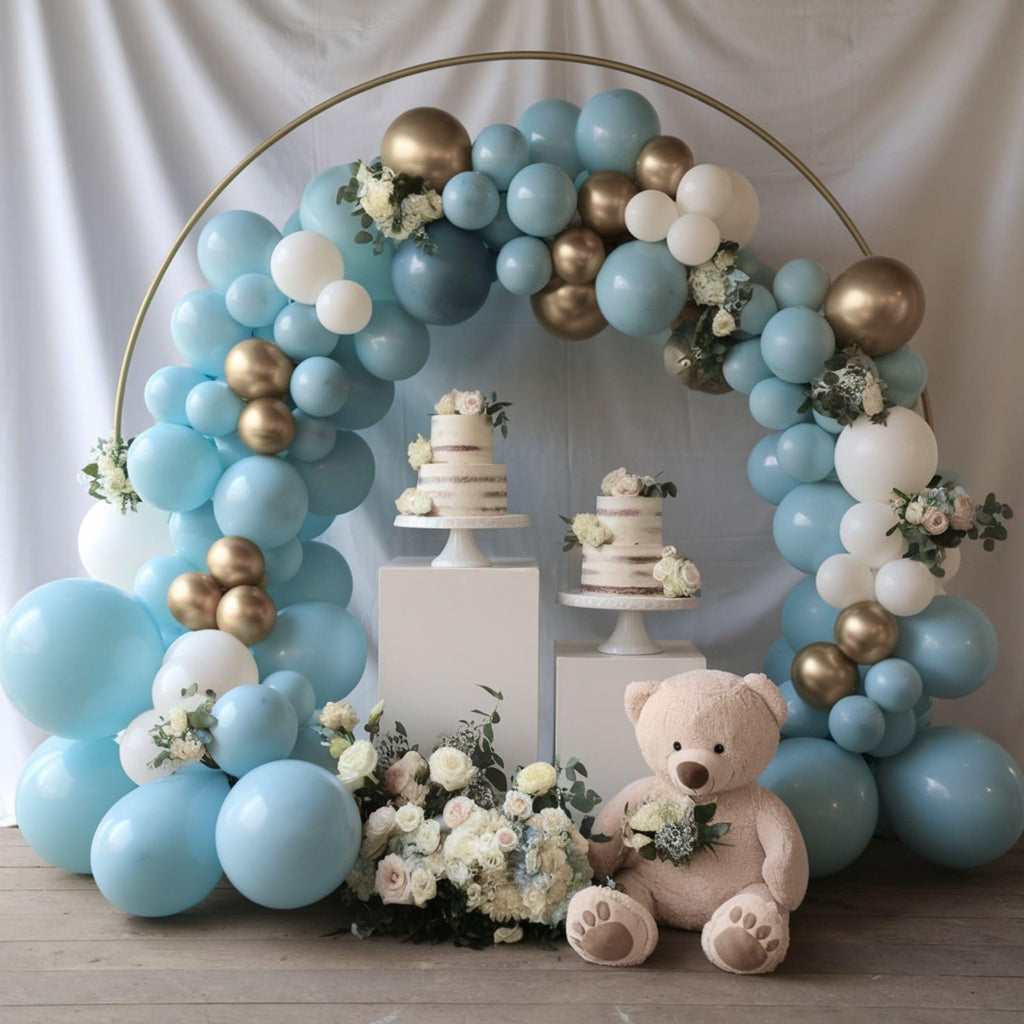 Balloon arch with dusty blue, white, and gold balloons framing two tiered cakes and a teddy bear.