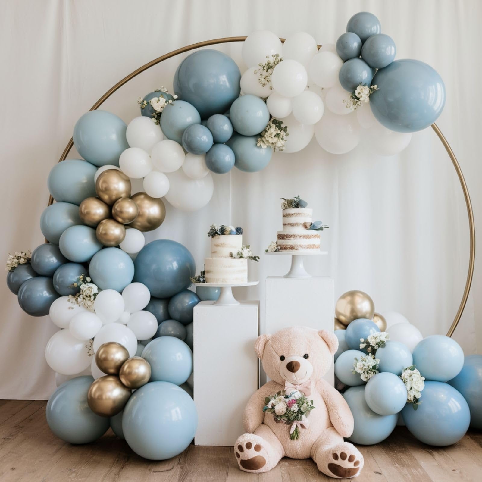 Dusty blue, gold, and white balloon arch with two small cakes and a teddy bear holding flowers