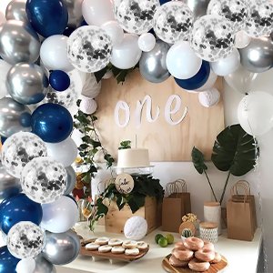 Party balloons in navy blue, pearl white, metallic silver, and silver confetti arranged around a wooden sign that says 'one', with a cake and donuts on a table.