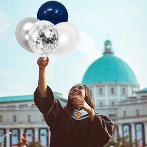 Graduating student in cap and gown holding a cluster of navy blue, pearl white, silver, and confetti balloons outdoors.