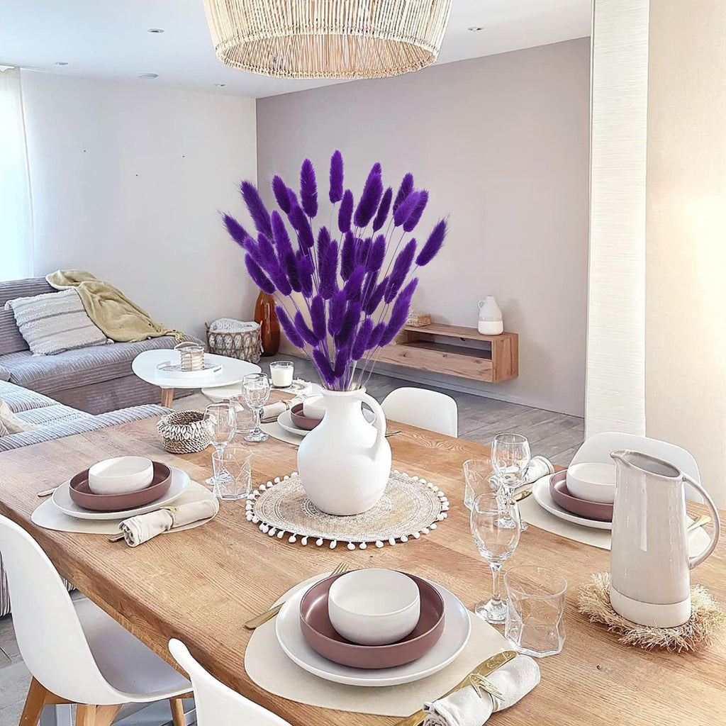 A dining table set for a meal with a vase of purple bunny tail grass as a centerpiece.