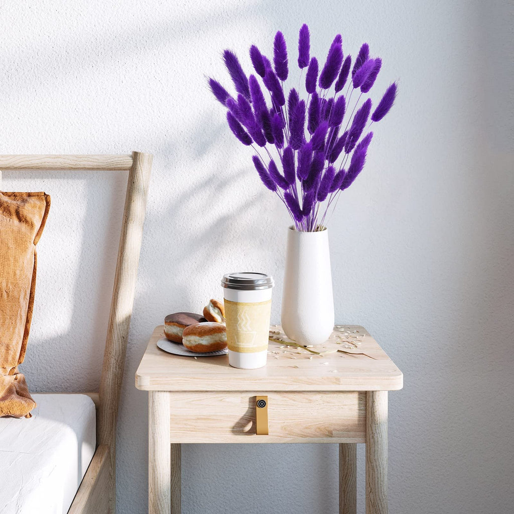 Purple bunny tail grass in a white vase on a wooden nightstand with donuts and coffee