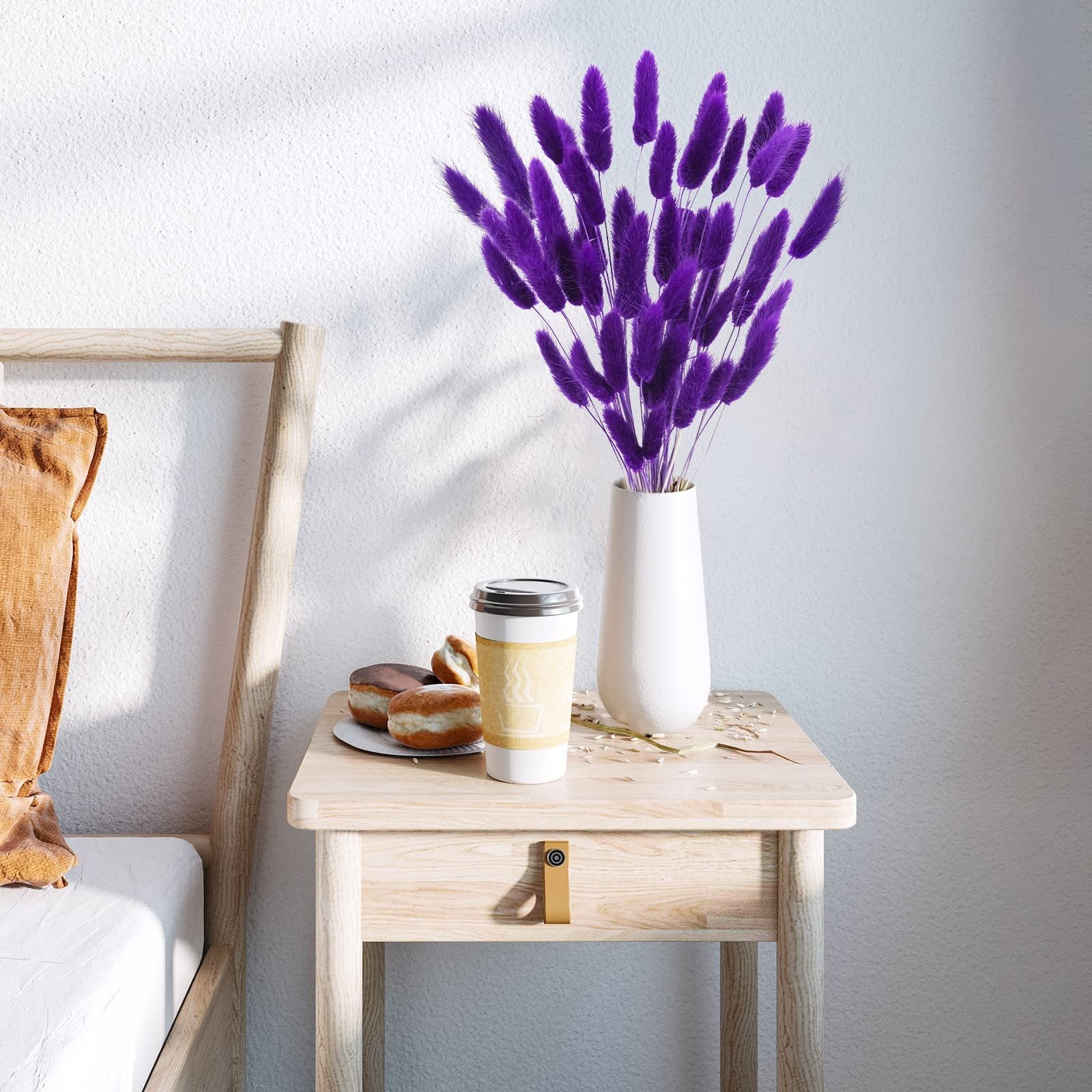 Purple bunny tail grass in a white vase on a wooden nightstand with donuts and coffee