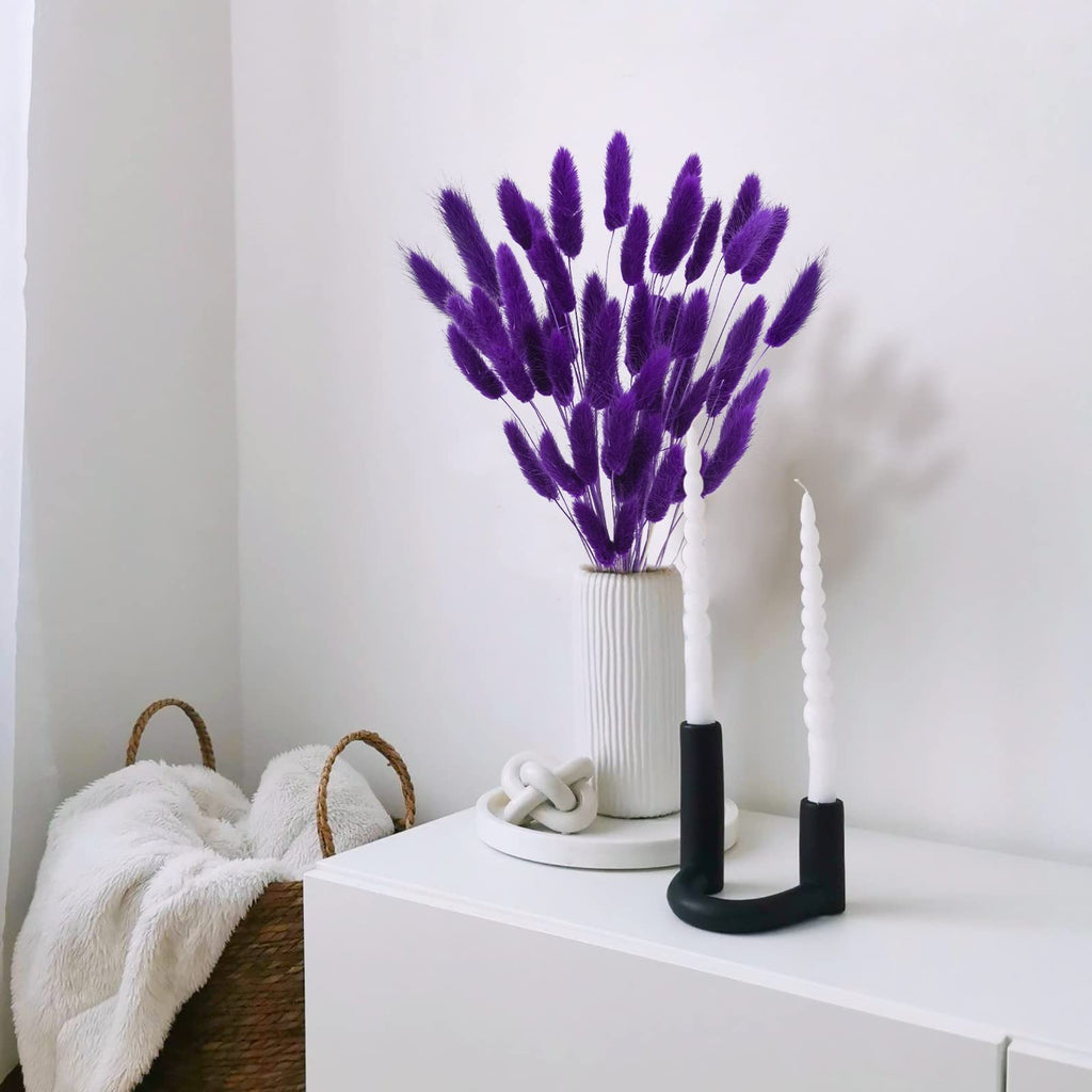 Purple bunny tail grass in a white vase with two white candles in a black holder on a white surface.